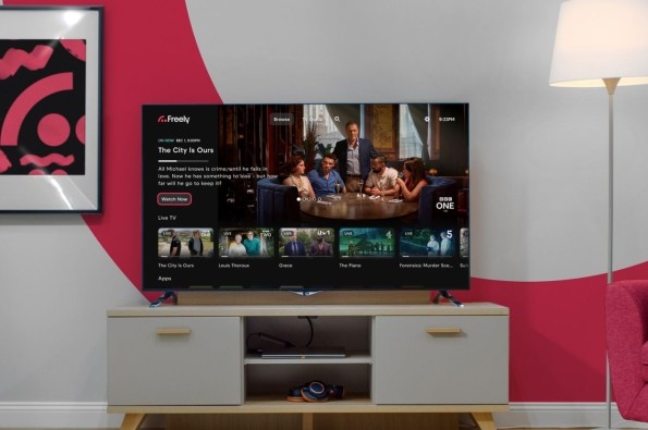 A lounge with a TV on a TV stand with a lamp next to it and the screen displaying the Freely user interface